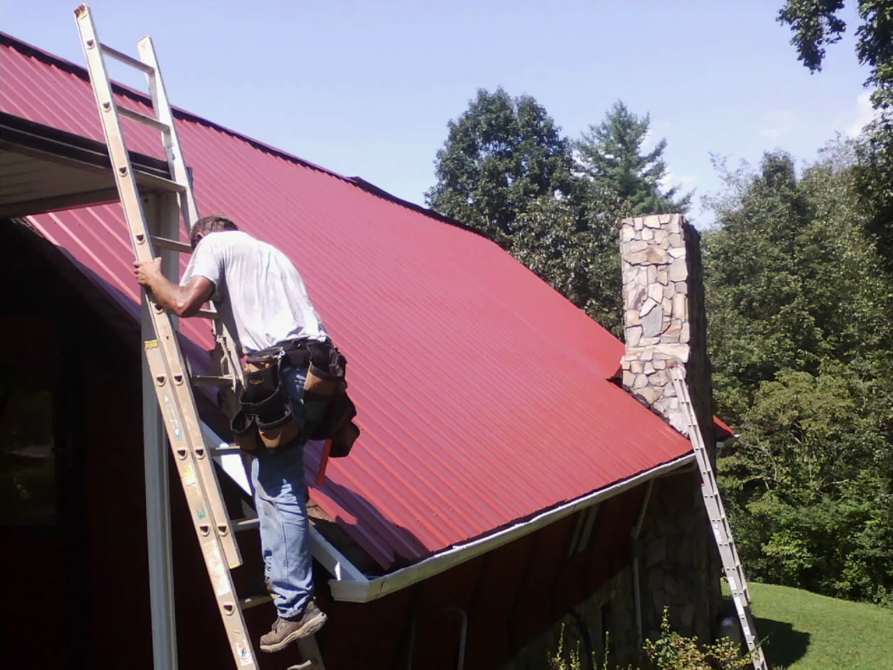 Roofer on a ladder beside a red metal roof and stone chimney — Soffit & Fascia Repair by Roofing Brothers in Roselle